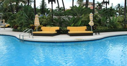 A large swimming pool with crystal clear blue water. The pool is surrounded by lush greenery and palm trees creating a tropical atmosphere. There are two yellow lounge chairs with orange and white striped umbrellas on either side of the pool providing shade for the lounge chairs. In the background there is a building with a red roof and white walls. The sky is blue and the overall mood of the image is peaceful and relaxing.