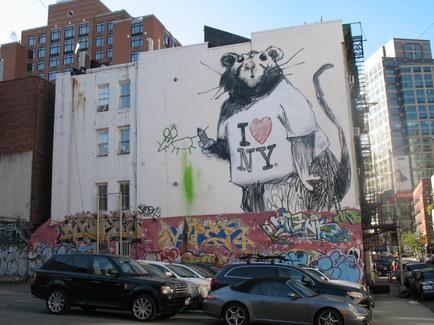 A large mural on the side of a building in New York City. The mural is a large black and white drawing of a rat with the words "I Love NY" written above it. The rat is holding a heart in its paws and is standing on its hind legs. The background of the mural is filled with colorful graffiti and there are several cars parked in front of it. On the right side of the image there are tall buildings with glass windows. The sky is blue and the weather appears to be sunny.