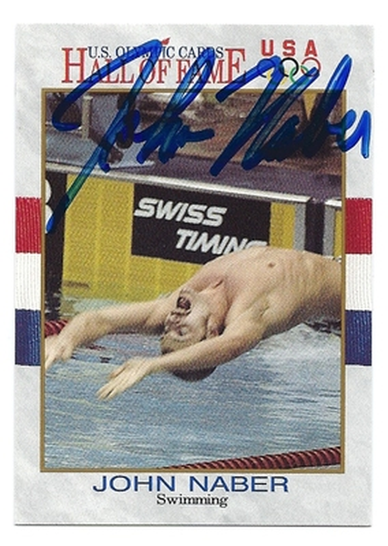 A photograph of John saber a professional swimmer from the United States. He is shown in mid-air with his arms stretched out to the sides and his body slightly tilted downwards. His head is tilted back and his eyes are closed as if he is in the middle of a dive. The background is blurred but it appears to be a swimming pool with a sign that reads "Swiss Timing" and "U.S. Olympic Games USA Hall of Fame." The photograph is signed by John nabbed in blue ink.