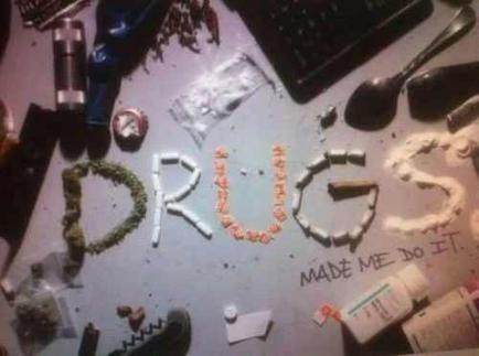 A desk with various items scattered on it. In the center of the desk there is a large word "DRUGS" written in white chalk. The word is made up of small orange and white pills with the words "Made me do it" written below it in a cursive font. Surrounding the word are various items such as a laptop a spoon a bottle of pills and a few other miscellaneous items. The desk appears to be cluttered and disorganized.