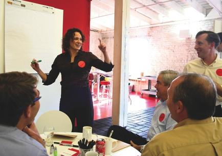 A group of people sitting around a table in a meeting room with a whiteboard in the background. There are six people in the room three men and three women all of whom are engaged in a conversation. The woman in the center of the room is standing in front of the whiteboard and is wearing a black blouse with red polka dots on it. She is holding a green marker and appears to be giving a presentation to the group. The man on the right side of the image is smiling and gesturing with his hand as he speaks. The other three men are sitting around the table listening attentively to the woman. The room has a brick wall and a large window on the left side allowing natural light to enter.