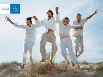 Four people two men and two women jumping in the air with their arms stretched out wide. They are all wearing white clothes and appear to be in a celebratory mood. The background shows a sandy beach with tall grass and a clear blue sky. The image also has a logo that reads "Live your life" in the top left corner. The overall mood of the image is joyful and carefree.