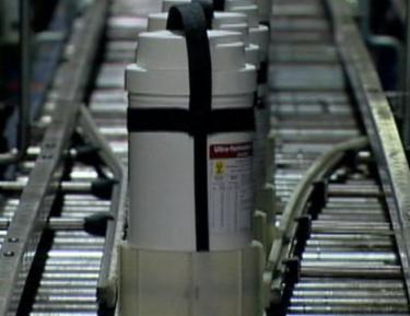 A conveyor belt in a factory or warehouse. The belt is made of metal and has a white plastic container with a black strap attached to it. The container has a label on it with a yellow star and the words "Vacuum Cleaner" written in black. The label also has some information about the product such as its size capacity and other specifications. The background is blurred but it appears to be a factory setting with other conveyor belts visible.