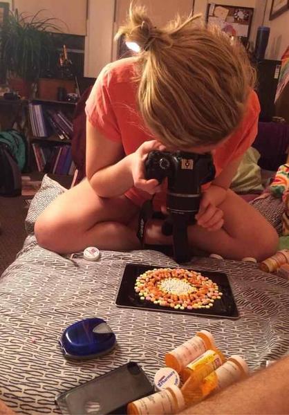A young woman sitting on a bed with her back to the camera. She is wearing an orange t-shirt and has blonde hair tied up in a messy bun. The woman is holding the camera with both hands and appears to be taking a photo of a plate of colorful candies on the bed. There are several prescription bottles scattered around the bed including a blue pill bottle a black phone case and a black tray with more candies. In the background there is a bookshelf and a plant.