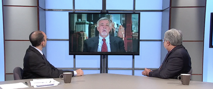 Two men sitting at a desk in a news studio engaged in a conversation. They are both wearing suits and appear to be in the middle of a news broadcast. On the desk there are two coffee mugs and some papers. In the background there is a large screen displaying a man in a suit and tie who appears to be a news anchor. The man in the suit is looking at the screen with a serious expression on his face. The room has a gray wall and a large window with a view of a city skyline.