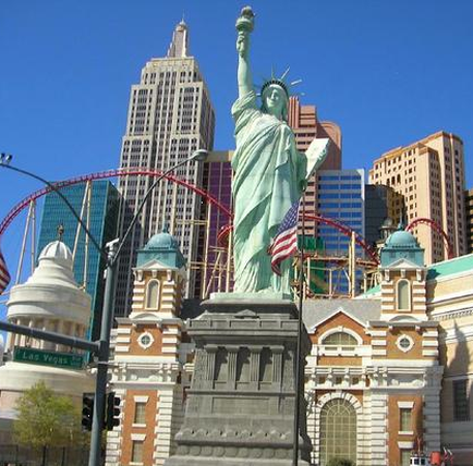 The Statue of Liberty in Las Vegas Nevada. The statue is standing on a pedestal and is holding an American flag in her right hand. She is facing towards the right side of the image. In the background there are several tall buildings and a roller coaster. The sky is blue and the weather appears to be sunny.