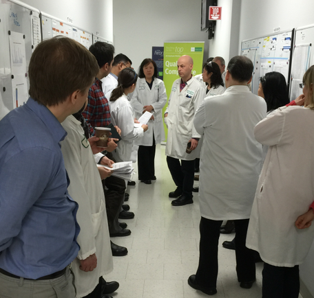 A group of people standing in a circle around a whiteboard in a classroom. They are all wearing white lab coats and appear to be engaged in a discussion or presentation. The people are of different ages and genders and they are all looking at the whiteboard with interest. Some of them are holding papers or documents while others are looking down at the board. There is a green banner in the background with writing on it. The room appears to be well-lit with natural light coming in from the windows.