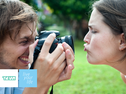 A man and a woman in a park. The man is holding a camera and taking a picture of the woman with it. The woman is kissing the man on the cheek. They are both smiling and appear to be enjoying the moment. The background is blurred but it appears to be a grassy area with trees and bushes. The image also has a logo that reads "tea" and the words "live your life".
