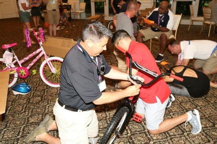 A group of people gathered in a room with a carpeted floor. In the center of the image there is a man wearing a black polo shirt and khaki pants kneeling on the floor and fixing a pink bicycle. He is holding the handlebars of the bicycle with both hands and appears to be in the process of fixing it. To his right there are two men one wearing a red shirt and the other wearing a white shirt who are kneeling down and looking at the man fixing the bike. Behind them another man is sitting on the ground and there are several other people in the background some of whom are also working on the bicycle. There are also several other bicycles and helmets scattered around the room.