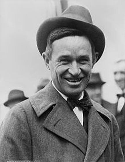 A black and white photograph of a man who appears to be in his late twenties or early thirties. He is wearing a suit and a hat and is smiling widely. He has short dark hair and is looking directly at the camera. In the background there are other men wearing hats suggesting that the photo was taken at an outdoor event. The man's expression is one of joy and contentment.