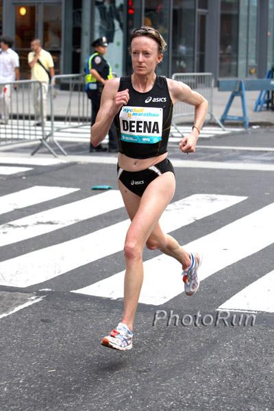 A woman running on a street. She is wearing a black tank top with the word "Deena" written on it and black shorts. She has a pair of sunglasses on her head and is wearing orange and white running shoes. The woman appears to be in the middle of a race as she is running with determination and focus. The street is lined with buildings and there are a few people visible in the background. The image appears to have been taken during the day.