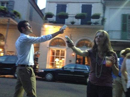 A man and a woman standing on a street at night. The man is on the left side of the image wearing a white shirt and khaki pants and is holding a white phone in his right hand. He is pointing towards the woman with his left hand as if he is about to take a picture or video call. The woman is standing next to him holding a drink in her hand. She is wearing a purple shirt and has long blonde hair. In the background there are other people walking on the sidewalk and a building with a balcony and potted plants. There is also a black car parked on the street. The sky is dark and the street is lit up with streetlights.