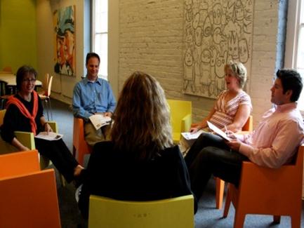 A group of five people sitting in a circle in a room with orange chairs. They are engaged in a conversation and appear to be engaged in some kind of activity. The room has a white brick wall with a large mural hanging on it and there are two large windows on the right side of the image. The people in the circle are of different ages and genders and they are all holding papers and pens in their hands. They seem to be of different ethnicity's and are dressed in casual clothing. The overall atmosphere of the room is relaxed and casual.