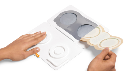 A pair of hands holding a white plastic tray with three circular compartments. The compartments are divided into three sections each with a different color - gray white and beige. The tray appears to be open as there is a small orange tag attached to one of the compartments with a label on it. The background is plain white.
