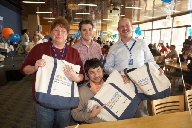 Four people three men and one woman standing in a large room with tables and chairs. They are all smiling and holding large blue and white bags with the word "EXALGO" written on them. The woman on the left is wearing a red shirt and glasses the man in the middle is wearing an orange shirt and the man on the right is wearing glasses and a blue lanyard. The room is decorated with blue and orange balloons and there are other people in the background. It appears to be an event or gathering.