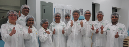 A group of nine people all wearing white lab coats and hairnets standing in a room with a whiteboard in the background. They are all smiling and giving a thumbs up with their thumbs up. The people appear to be happy and proud of their work.