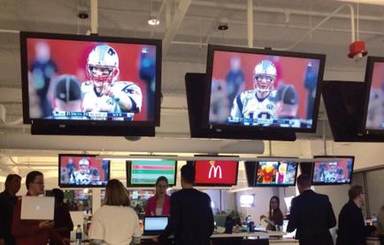 A McDonald's restaurant with multiple large screens hanging from the ceiling. The screens display a football player wearing a white helmet and holding a football. The player appears to be in the middle of a game as there are other players visible in the background. In the foreground there are several people standing in front of the screens some of them are looking at their laptops. The McDonald's logo is visible on the right side of the image.