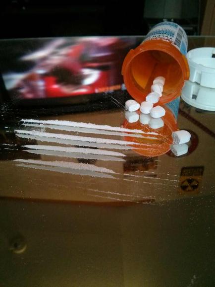 An open prescription bottle with white pills spilling out of it onto a brown surface. The bottle is orange and white in color and has a label on it. The pills are scattered around the bottle some of them are spilling out and some are lying on the surface. In the background there is a TV screen with a red and black image of a person's face on it suggesting that the image was taken in a dimly lit room.