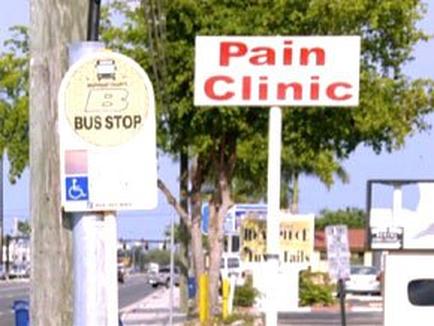 A bus stop sign on the side of a street. The sign is rectangular in shape and has a white background with black text. On the left side of the sign there is a yellow circle with the words "BUS STOP" written in black letters. Next to the circle there are two smaller white signs with red text that read "Pain Clinic" and "Bus Stop". On the right side the same sign is white with black lettering. The street is lined with trees and there are cars parked on the sidewalk. The sky is blue and the weather appears to be sunny.
