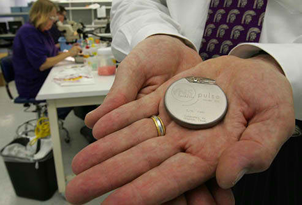 A person's hands holding a small silver coin. The coin appears to be made of metal and has the word "Pulse" written on it in black letters. The person is wearing a white shirt and a purple tie. In the background there is a desk with a computer and other laboratory equipment. A woman is sitting at the desk working on a project. The overall atmosphere of the image is professional and focused.
