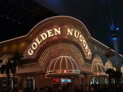 The entrance of the Golden Nugget Casino in Las Vegas Nevada. The building is a large ornate structure with a curved archway and a sign that reads "Golden Nugget" in large golden letters. The entrance is lit up with bright lights creating a warm glow. The sky is dark and there are palm trees and other buildings in the background. People can be seen walking around the entrance and a street lamp is visible on the right side of the image. The overall atmosphere is lively and bustling.