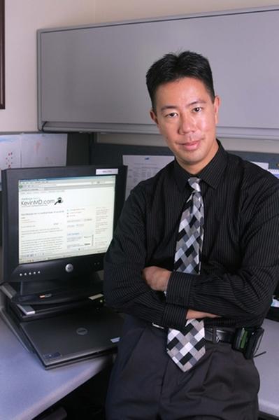 A young man sitting at a desk in an office. He is wearing a black shirt with a checkered tie and has his arms crossed in front of him. He has a smile on his face and is looking directly at the camera. Behind him there is a computer monitor and a laptop on the desk. The desk is cluttered with papers and other office supplies. The man appears to be in his late twenties or early thirties.