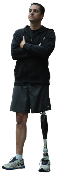 A young man standing with his arms crossed in front of a brick building. He is wearing a black goodie black shorts and white sneakers. He has a prosthetic leg that is attached to his left leg. The man is looking up towards the sky with a serious expression on his face. The background is blurred but it appears to be an outdoor setting with plants and flowers.