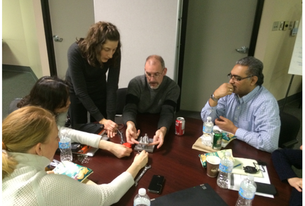 A group of five people sitting around a wooden table in a meeting room. There are four people in the image three men and two women all of whom appear to be engaged in a discussion. The table is covered with a brown tablecloth and there are various items on it including water bottles a laptop a can of soda and a notebook. The woman on the left is standing and appears to be explaining something to the group while the man on the right is sitting and looking at the laptop screen. The other three people are sitting around the table and one of them is holding a pen and writing on a piece of paper. The room has a whiteboard and a door in the background.