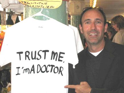 A man standing in front of a white t-shirt with the words "Trust me I'm a doctor" written on it in black letters. The man is pointing at the shirt with his right hand and is smiling at the camera. He is wearing a black suit and has a pair of sunglasses on his head. Behind him there are other people in the background suggesting that he is at a market or market stall. The shirt appears to be a doctor's office or a clothing store.