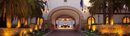 The entrance of a luxurious hotel named Bacara. The entrance is a large archway with a white building in the background. The building has multiple arched windows and a red tiled roof. The archway is surrounded by palm trees and other tropical plants. On either side of the archway there is a stone wall with the word "Bacara" written on it. The sky is dark indicating that it is either dusk or early morning. The overall atmosphere of the image is peaceful and inviting.