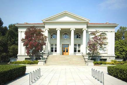 A large two-story building with a white exterior and a red roof. The building has a pediment on the top which is supported by four columns. The front entrance has a large wooden door with a glass panel in the center. There are two columns on either side of the door and a set of stairs leading up to the entrance. The entrance is flanked by two large windows on the second floor and a small balcony on the third floor. The building is surrounded by a well-manicured garden with neatly trimmed bushes and trees. The sky is blue and there are a few clouds in the sky. In front of the building there is a pathway leading to the front door. The overall atmosphere of the image is peaceful and elegant.