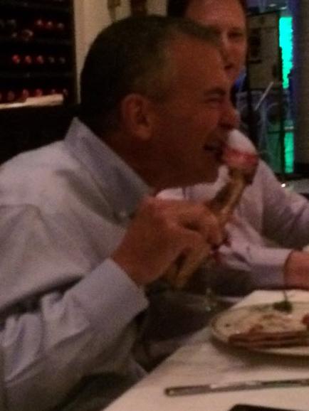 Two men sitting at a table in a restaurant. The man on the left is wearing a light blue collared shirt and is holding a glass of wine in his hand. He is smiling and appears to be enjoying his drink. On the table in front of him there is a plate of food and a knife. The background is blurred but it seems like the restaurant is dimly lit.