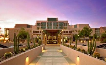 A modern mufti-story building with a large entrance. The building is made of light-colored concrete and has multiple levels with large windows and balconies. The entrance is flanked by two large arched windows on either side. The sky is a beautiful pink and purple color indicating that it is either sunrise or sunset. In front of the building there is a long walkway with a variety of cacti and succulents planted along the sides. The walkway is lined with small lights creating a warm and inviting atmosphere. On the right side of the image there are other buildings and trees adding to the urban feel of the scene.