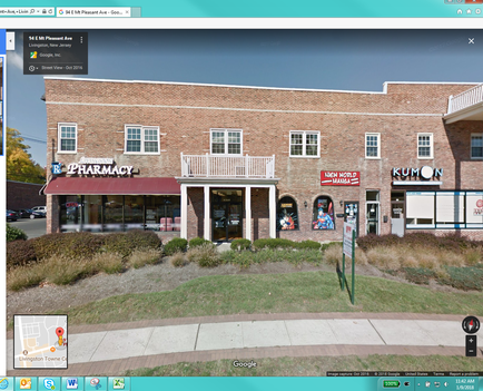 A street view of a brick building with a pharmacy on the left side. The building has a red awning with the word "Pharmacy" written on it in white letters. On the right side of the building there is a KUMON store with a sign that reads "KUMON". The building is located on a street corner with a sidewalk in front of it. There are trees and bushes surrounding the building. The sky is blue and there are a few clouds in the background.
