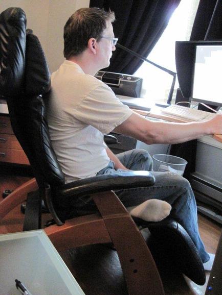 A man sitting in an office chair in front of a computer monitor. He is wearing a white t-shirt blue jeans and white socks. He has short brown hair and is wearing glasses. The desk is cluttered with a keyboard mouse and other office supplies. There is a window with black curtains in the background. The man appears to be focused on his work.