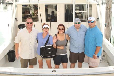 A group of five people standing on the deck of a boat. There are six people in the group four men and three women all wearing sunglasses and hats. They are all smiling and posing for the photo. The boat is docked at a marina with other boats in the background. The woman in the center is holding a black tote bag and making a peace sign with her hand. The man on the left is wearing a beige shirt and khaki shorts the man in the middle is wearing sunglasses and the woman on the right is wearing an orange shirt and a baseball cap. All of them are wearing casual summer clothes.