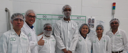 A group of nine people nine men and nine women standing in a laboratory or laboratory setting. They are all wearing white lab coats and hairnets and are standing in front of a whiteboard with the words "DO CHEF ACT" written on it. The men are smiling and giving a thumbs up indicating that they are happy and proud of their work. The women are also smiling and looking at the camera. They appear to be of different ages and ethnicity's. The background of the image is a white wall with pipes and other laboratory equipment.