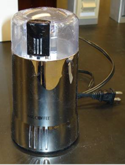 A coffee grinder on a kitchen countertype. The grinder is made of stainless steel and has a cylindrical shape with a transparent lid on top. The lid has a black knob on the top for adjusting the temperature. There is a power cord attached to the grinder which is plugged into a power socket on the right side of the image. The brand name "NRL COFFEE" is visible on the front of the grinders.