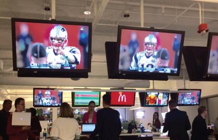 A large room with multiple large screens hanging from the ceiling. The screens are displaying a football game between the New England Patriots and the Philadelphia Eagles. In the center of the room there is a McDonald's logo and a group of people standing in front of the screens. The people in the room appear to be engaged in the game with some looking at the screens and others looking at their laptops. The room has a high ceiling with exposed pipes and outwork.