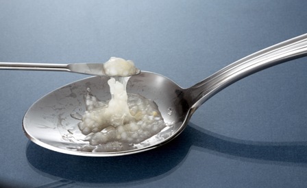 A silver spoon with a small amount of white substance likely an opioid or another drug on it. A smaller metal scoop appears to be stirring or scooping the white substance from the spoon.  The spoon is resting on a blue surface.