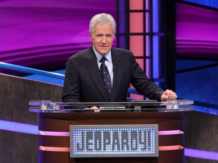 A man standing behind a desk with a sign that reads "Jeopardy!" The man is wearing a dark suit and tie and appears to be in the middle of a jeopardy game. He is standing in front of a stage with a blue and purple background and a large screen behind him. The man has gray hair and is looking directly at the camera with a serious expression on his face. On the desk there is a nameplate with the word "jeopardy!" written on it.