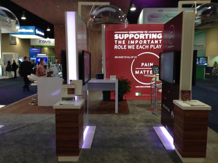 A trade show booth with a red banner that reads "Supporting the important role we each play" and "Pain Matte". The booth is located in a large exhibition hall with other booths and people in the background. There are two large screens on either side of the booth and a desk in front of it with a computer monitor and other items on it. The booth has a modern design with white walls and a wooden floor. The overall atmosphere of the exhibition hall is professional and inviting.