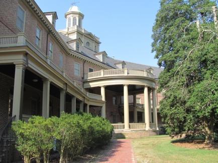 A large brick building with a clock tower on top. The building has multiple levels with columns and a balcony on the second floor. The balcony has a railing and there are trees and bushes surrounding it. The sky is blue and the weather appears to be sunny. There is a brick pathway leading up to the building and a grassy lawn on the right side of the image.