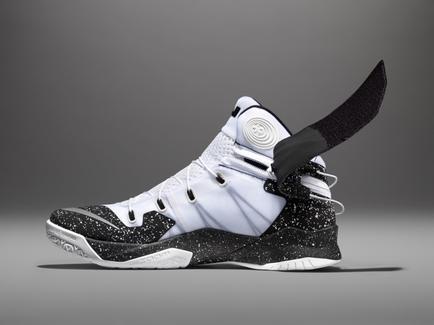 A photograph of a white and black basketball shoe. The shoe has a white upper with black accents and a black sole. The laces are white and the tongue of the shoe is black with a white logo in the center. The heel of the sneaker has a black strap that wraps around the ankle. The background is a plain grey color. The image is taken from a side angle looking up at the shoe.