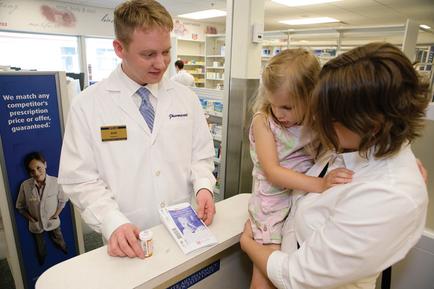 A young man in a white lab coat standing at a counter in a pharmacy. He is holding a prescription bottle and appears to be explaining something to a young girl who is standing next to him. The girl is wearing a pink dress and is looking at the man with a curious expression on her face. On the counter there is a sign that reads "We match any prescription price or offer guaranteed." In the background there are shelves with various medicines and other pharmacy equipment.