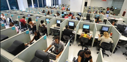 A large open-plan office space with multiple cubicles arranged in rows. The cubicles are arranged in a neat and orderly manner with people working on their computers. The people are of different ages genders and ethnicity's and they appear to be engaged in their work. The room has large windows on the far wall allowing natural light to enter. The floor is made of concrete and there are several desks and chairs scattered throughout the space. The overall atmosphere of the space is busy and productive.
