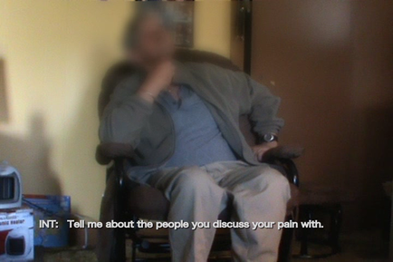 An elderly man sitting in a chair with his head in his hands. He is wearing a grey jacket and beige pants. The background is blurred but it appears to be a living room with a yellow wall and a wooden floor. On the right side of the image there is a TV and some other items scattered around the room. The man's face is not visible in the image. The text on the image reads "INT: Tell me about the people you discuss your pain with."