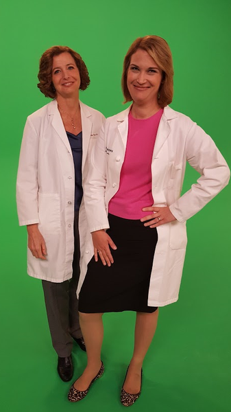 Two women standing side by side in front of a green background. They are both wearing white lab coats and are smiling at the camera. The woman on the left has short brown hair and is wearing a navy blue blouse and a black skirt. She is also wearing a pink top and black heels. Both women have their hands on their hips and appear to be posing for the photo.