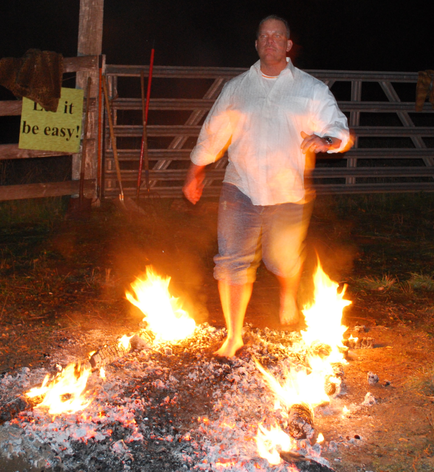 A man standing in front of a bonfire at night. He is wearing a white shirt and blue jeans and appears to be in the middle of a dance move. The bonfire is burning brightly with orange and yellow flames. The man is standing on a pile of rocks and there is a wooden fence in the background with a sign that reads "Let it be easy!" The sky is dark and the ground is covered in grass.
