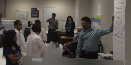 A group of people in a classroom setting. There are six people in the image three men and three women all wearing white lab coats. They are standing in front of a whiteboard and appear to be engaged in a discussion. The man in the center of the image is pointing to a poster on the whiteboard with his right hand and appears to be explaining something to the group. He is wearing a blue shirt and glasses and is gesturing with his left hand as he speaks. Behind him there are several other people standing and looking at the poster. On the left side of the room there is a desk with a computer monitor and other office supplies. The room is well-lit with natural light coming in from the windows.